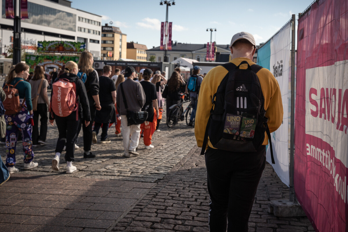 Keltaiseen takkiin ja reppuun pukeutunut henkilö kävelee mukulakivikadulla kohti ihmisjoukkoa ulkoilmatapahtumassa kaupunkialueella, taustalla näkyy rakennuksia ja banderolleja.