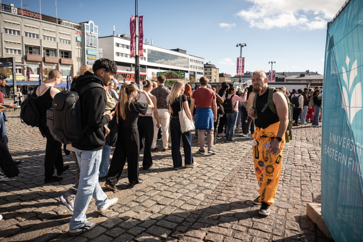 Ihmisjoukko seisoo jonossa kaupungin mukulakiviaukiolla. Oransseihin housuihin pukeutunut mies, jolla on laastareita, poseeraa oikealla olevan sinisen kyltin lähellä. Taustalla on moderneja rakennuksia ja aurinkoinen taivas.