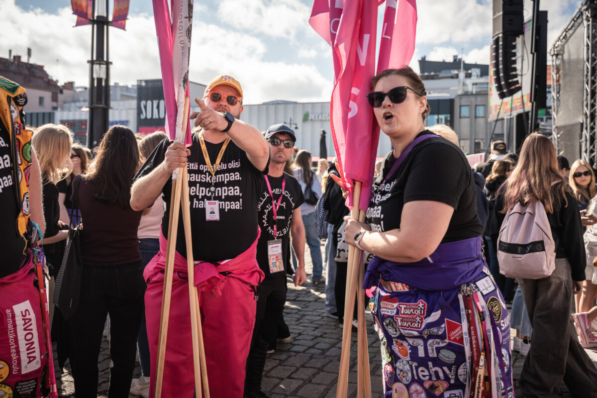Joukko värikkäisiin asuihin pukeutuneita ihmisiä pitelee vaaleanpunaisia lippuja ja banderolleja ulkoilmatapahtumassa. Kahdella edessä olevalla henkilöllä on aurinkolasit ja mustat paidat, kun taas taustalla näkyy muita ja kaupunkikuva osittain pilvisen taivaan alla.