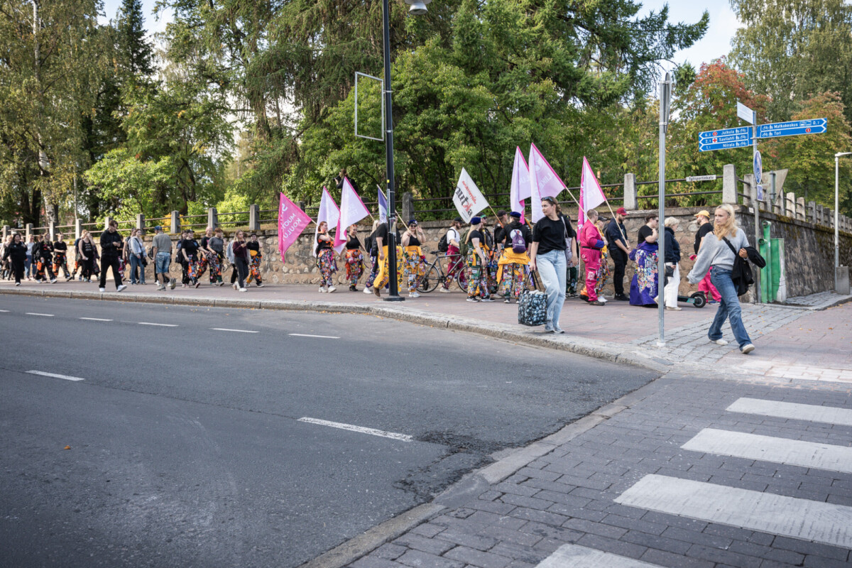 Joukko ihmisiä kävelee jalkakäytävällä vaaleanpunaiset liput ja banderollit kädessään ja osallistuu marssiin tai mielenosoitukseen. Kohtaus sijoittuu kaupunkialueelle, jossa näkyy puita, kivimuuri ja katukylttejä.