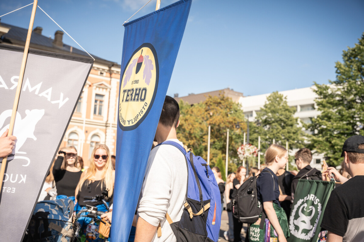 Joukko ihmisiä kokoontuu ulkona aurinkoisena päivänä, ja joillakin on kädessään suuria lippuja ja banderolleja, joista yksi on sininen, jossa lukee TERHO. Taustalla näkyy historiallisia rakennuksia ja vihreitä puita.