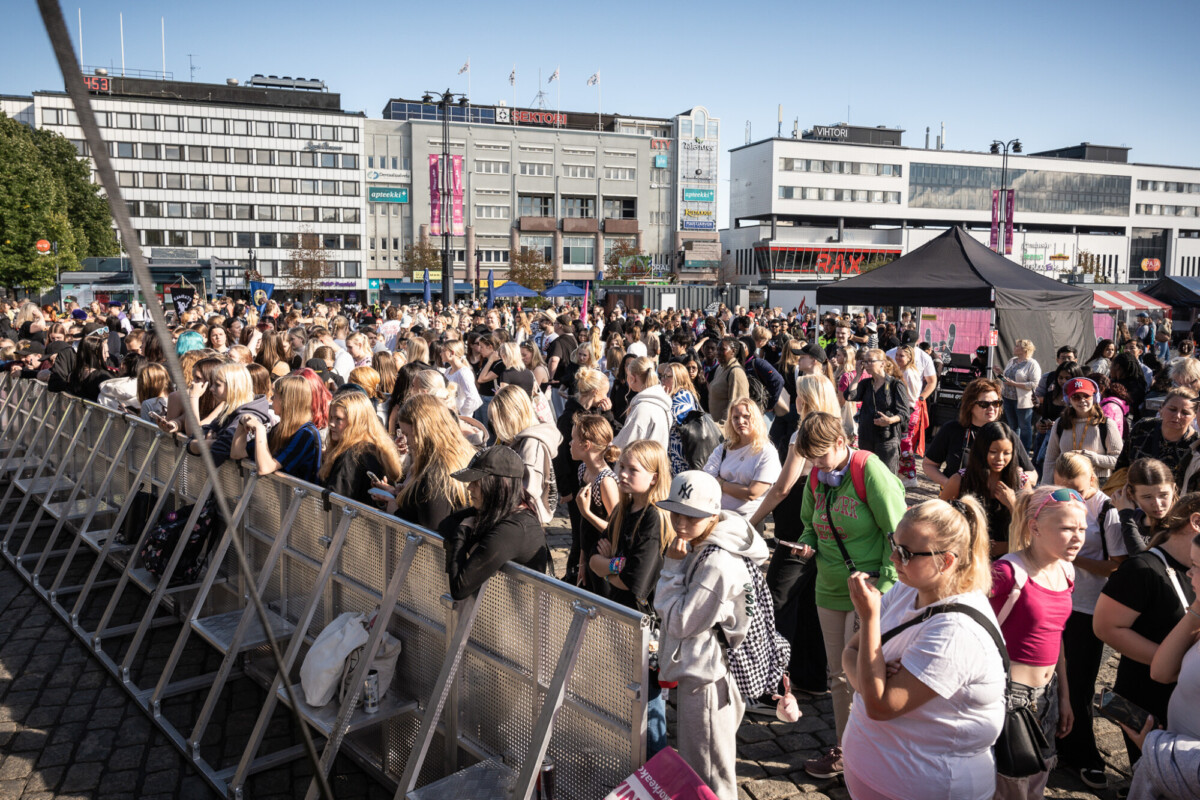 Suuri ihmisjoukko kokoontuu ulkona kaupungin aukiolla aurinkoisena päivänä, seisoo metalliesteiden takana ja taustalla näkyy rakennuksia ja telttoja.