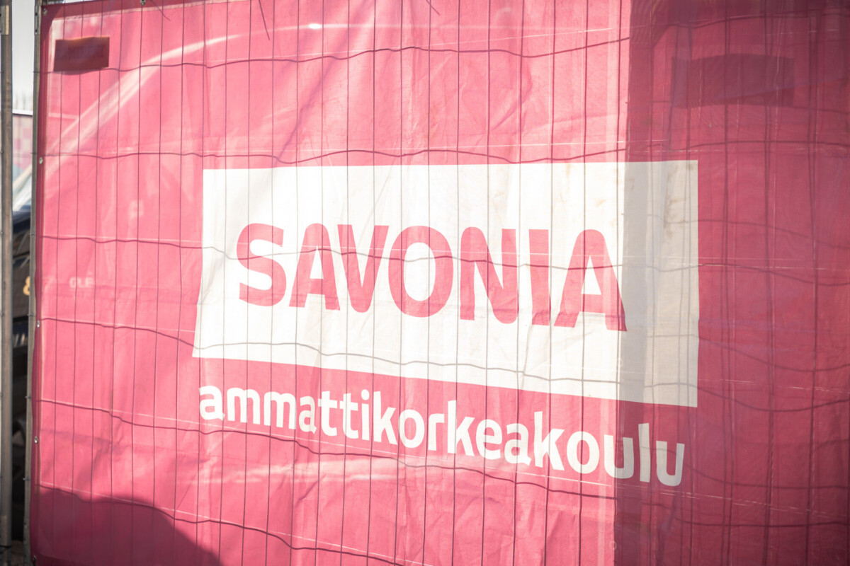 Metalliaitaan kiinnitetty vaaleanpunainen banderolli, jossa on valkoisella painettu teksti SAVONIA ammattikorkeakoulu. Auringonvalo heittää varjoja pintaan.