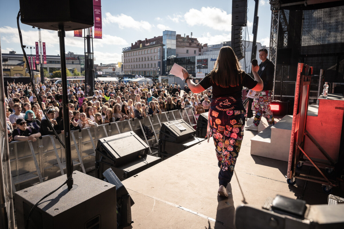 Henkilö, jolla on värikkäiden laastareiden peittämät housut, puhuu lavalla suurelle ulkoilmayleisölle aurinkoisena päivänä. Taustalla näkyy rakennuksia ja festivaalibanderolleja.