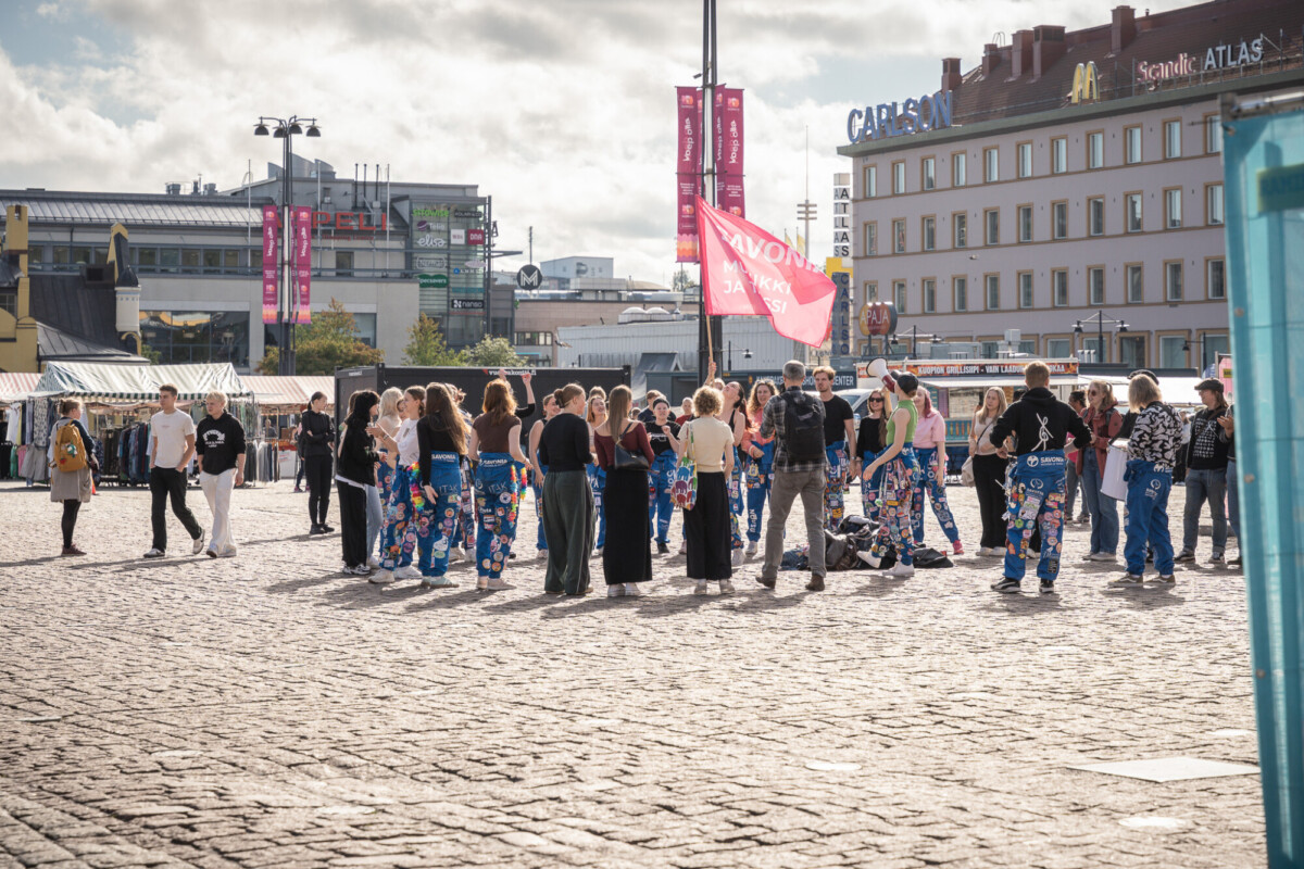 Joukko ihmisiä, joista monilla on värikkäät haalarit, seisoo mukulakiviaukiolla aurinkoisena päivänä. Taustalla näkyy rakennuksia, lippuja ja myyntikojuja.