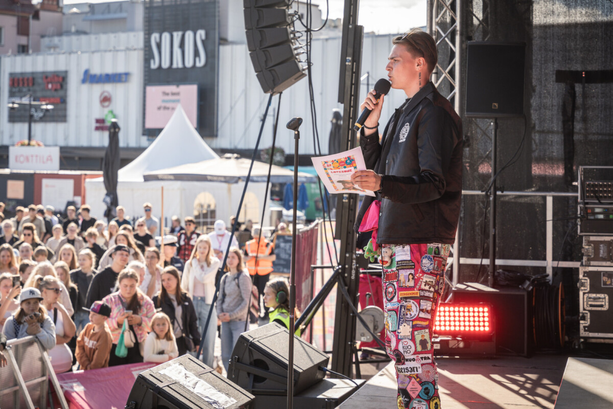Henkilö, jolla on kädessään mikrofoni ja värikäs vihkonen, seisoo lavalla ja puhuu suurelle ulkoilmayleisölle. Yleisö seuraa tarkkaavaisesti, ja taustalla näkyy kauppoja ja tapahtumateltoja.