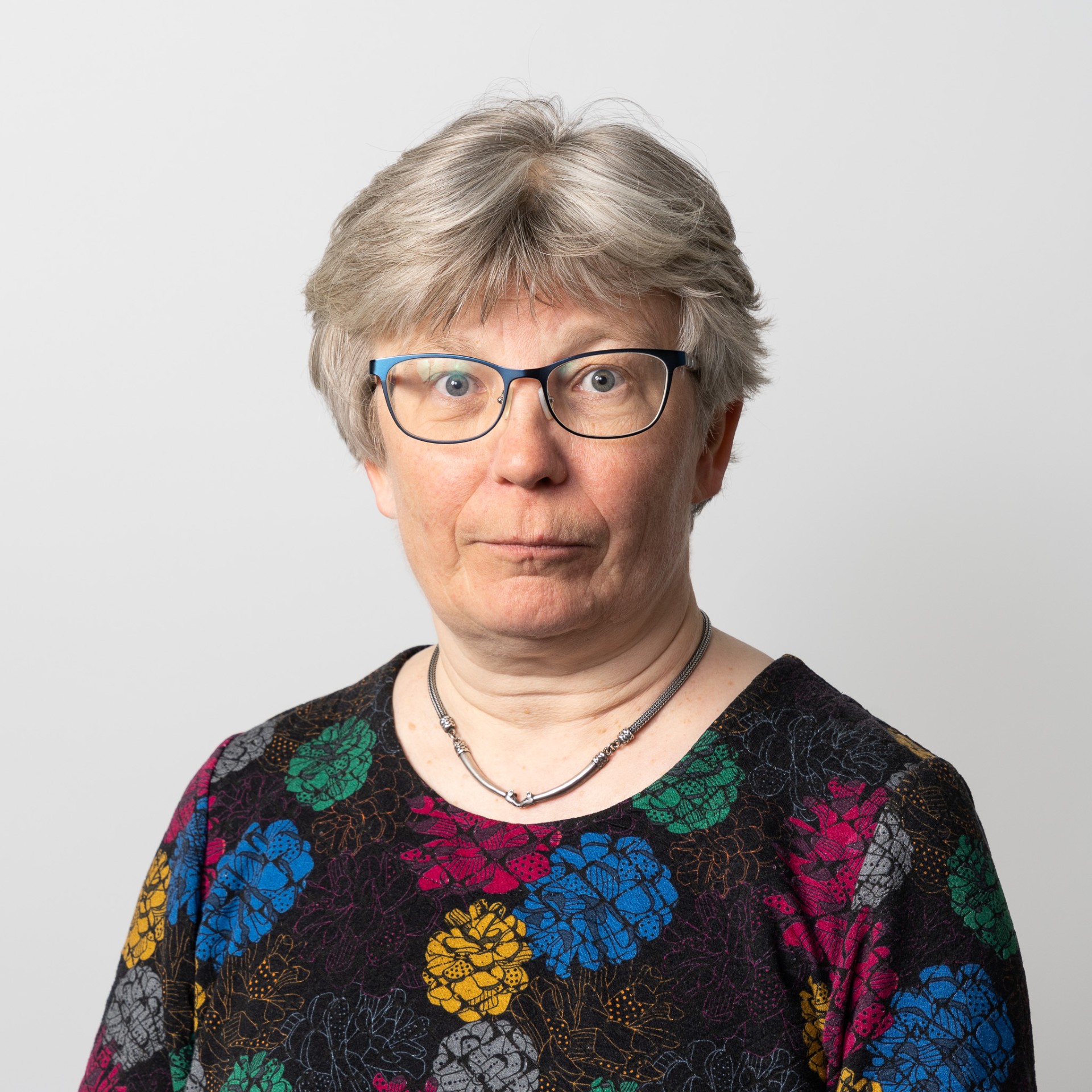 Keski-ikäinen henkilö, jolla on lyhyet harmaat hiukset ja silmälasit ja jolla on yllään värikäs kukkakuvioinen toppi ja kaulakoru, seisoo vaaleaa taustaa vasten ja katsoo suoraan kameraan neutraalilla ilmeellä.