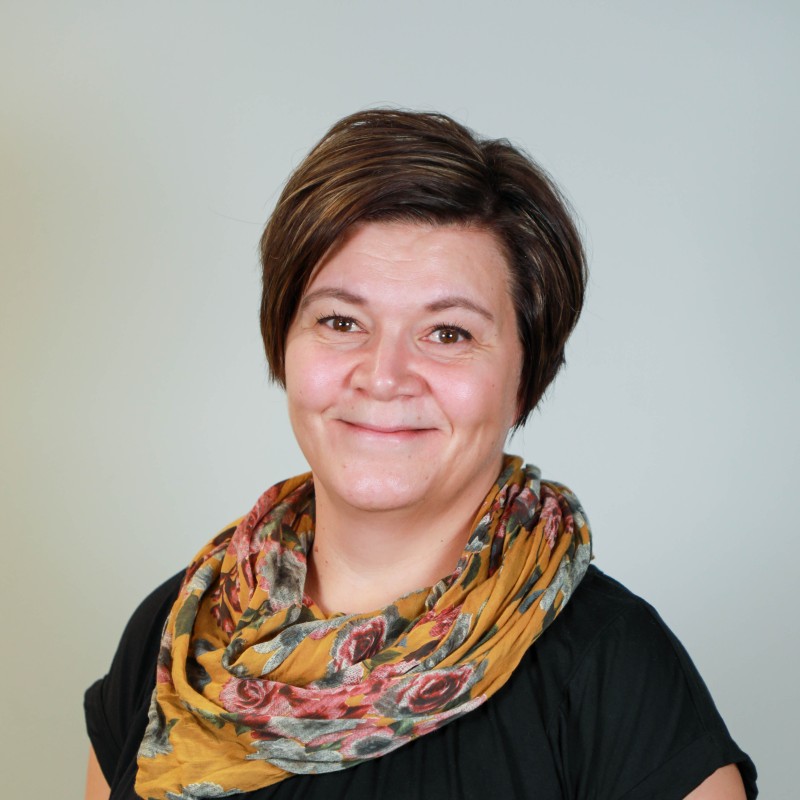 Lyhyet ruskeat hiukset ja kukkahuivi päässä oleva nainen hymyilee kameralle, päällään musta toppi, taustan ollessa vaalea.