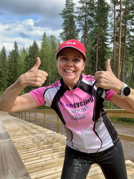 Hymyilevä nainen vaaleanpunaisessa ja mustassa pyöräilypaidassa ja punaisessa lippiksessä nostaa kaksi peukkua ylös metsäisellä puupolulla, jonka taustalla on korkeita puita ja pilvinen taivas.