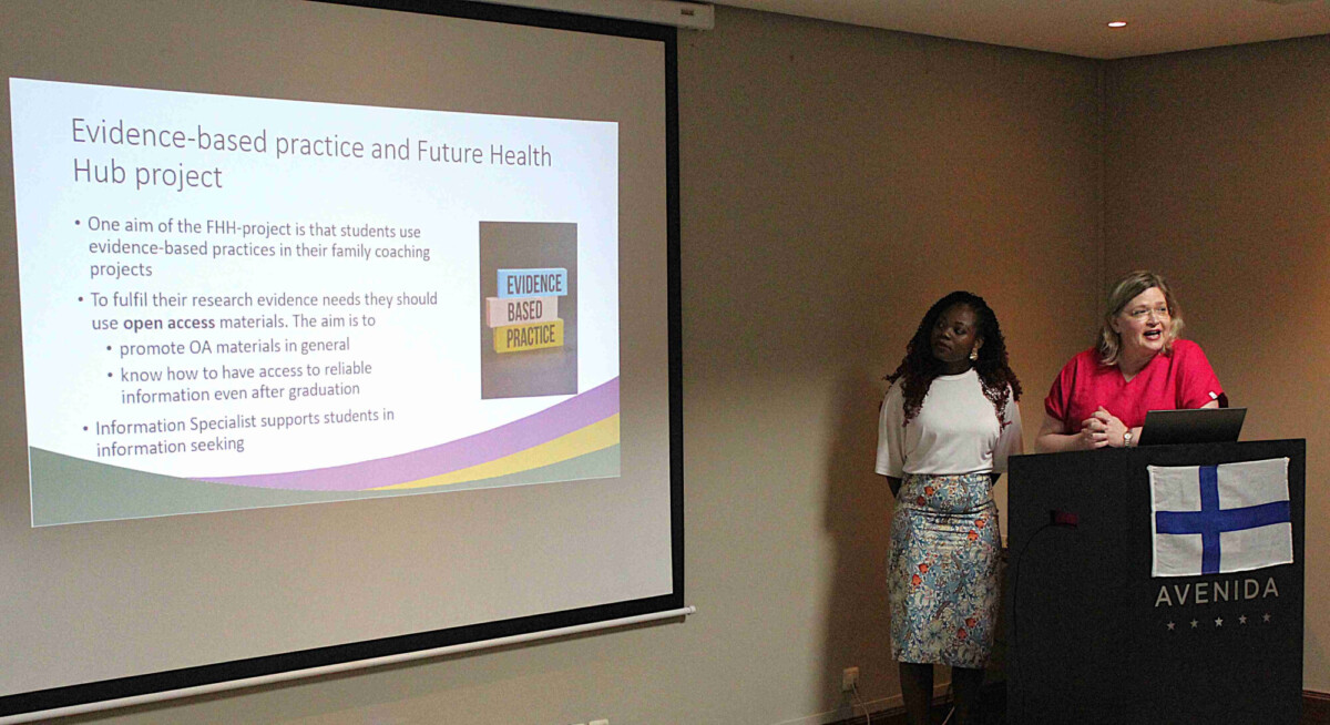 Kaksi naista seisoo kokoushuoneessa esitysnäytön vieressä, jossa näkyy dia, jonka otsikkona on näyttöön perustuva käytäntö ja Future Health Hub -hanke. Toinen nainen puhuu ja toinen kuuntelee.