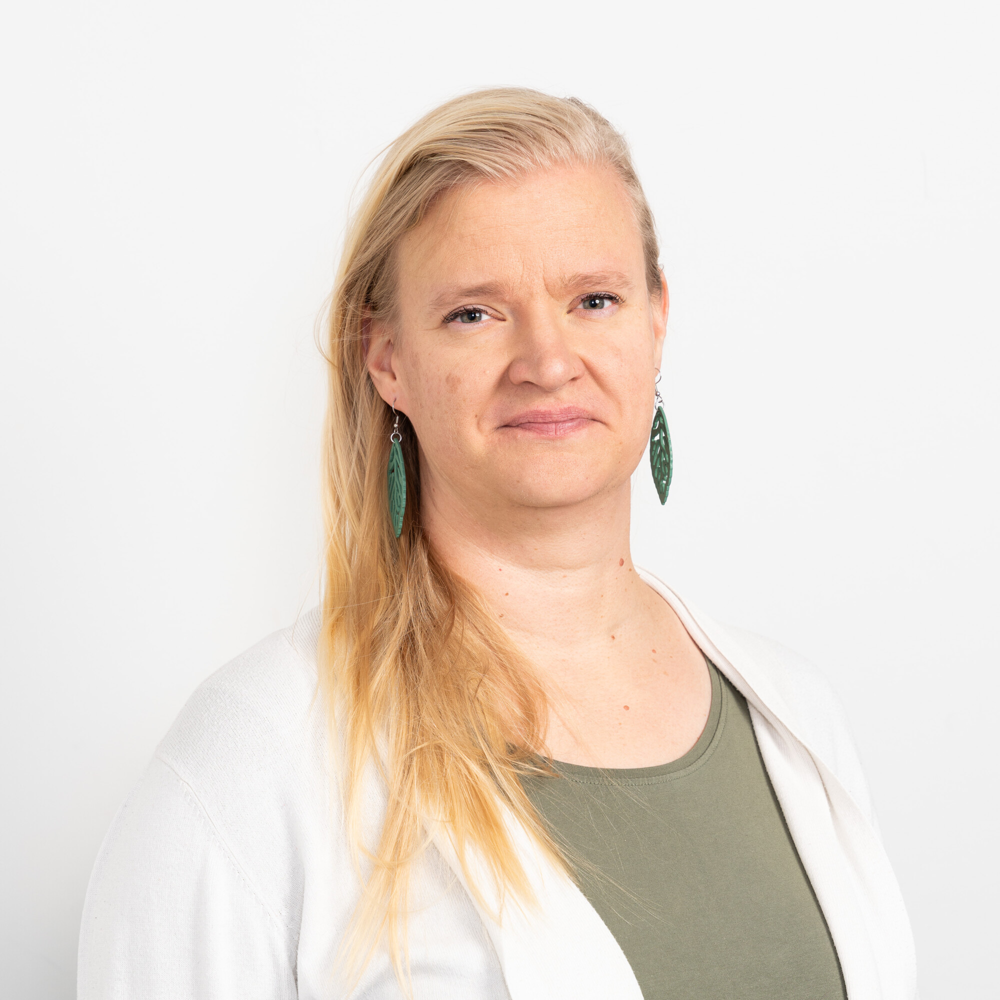 Pitkät vaaleat hiukset omaava henkilö, jolla on vihreät korvakorut, vihreä toppi ja valkoinen neuletakki, seisoo valkoista taustaa vasten ja katsoo suoraan kameraan neutraalilla ilmeellä.