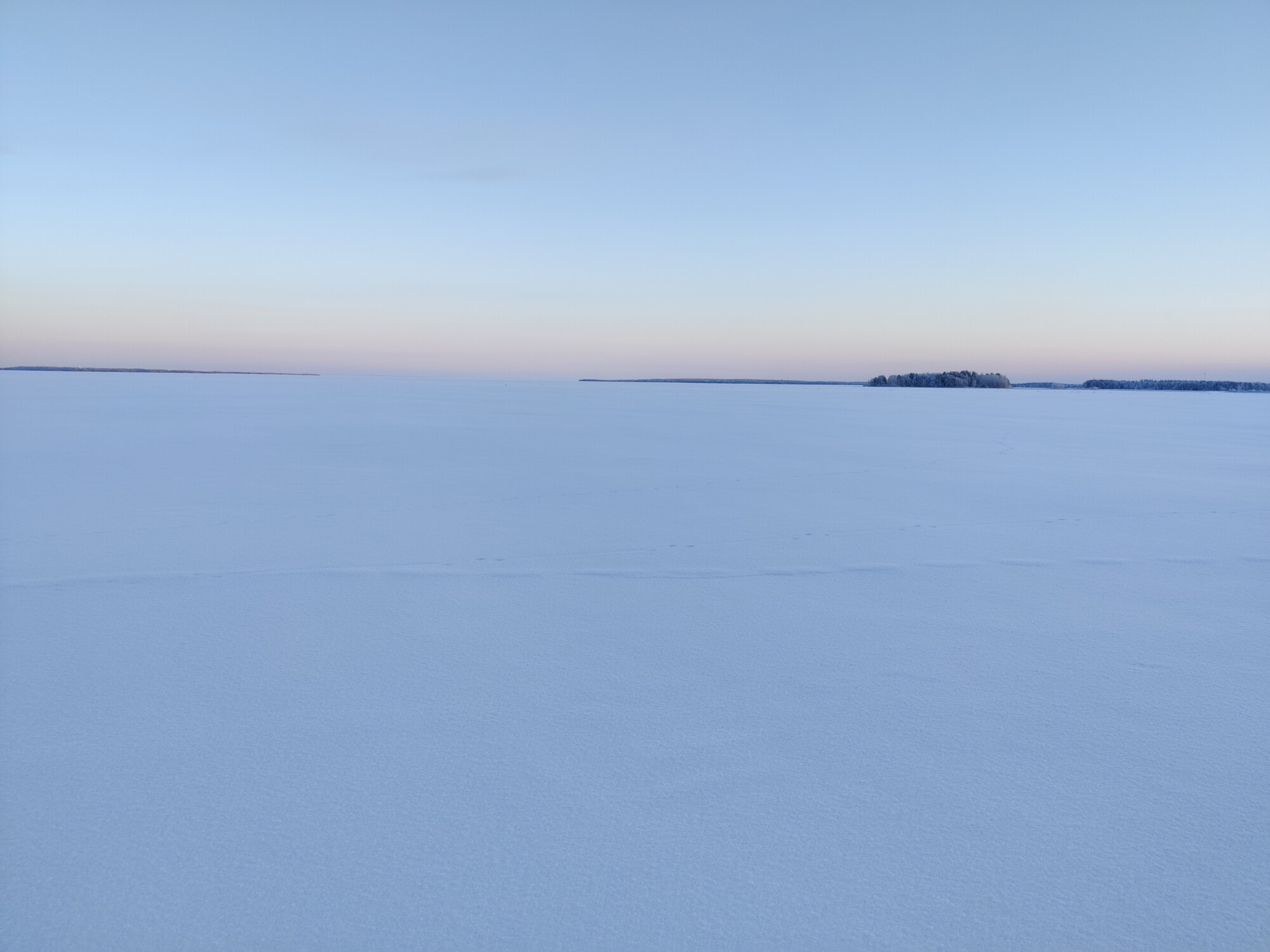 Auringon noustessa tai laskiessa vaaleansinisen ja vaaleanpunaisen taivaan alla levittäytyy laaja lumipeitteinen maisema, ja kaukana horisontissa näkyy hädin tuskin puiden reunustama maa.