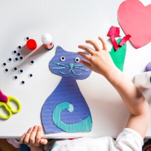 Lapsen kädet tekevät siniraidallista paperikissaa, jolla on kikkasilmät, ja ympärillä on askartelutarvikkeita, kuten sakset, liimaa, värillistä paperia ja maalipulloja valkoisella pöydällä.
