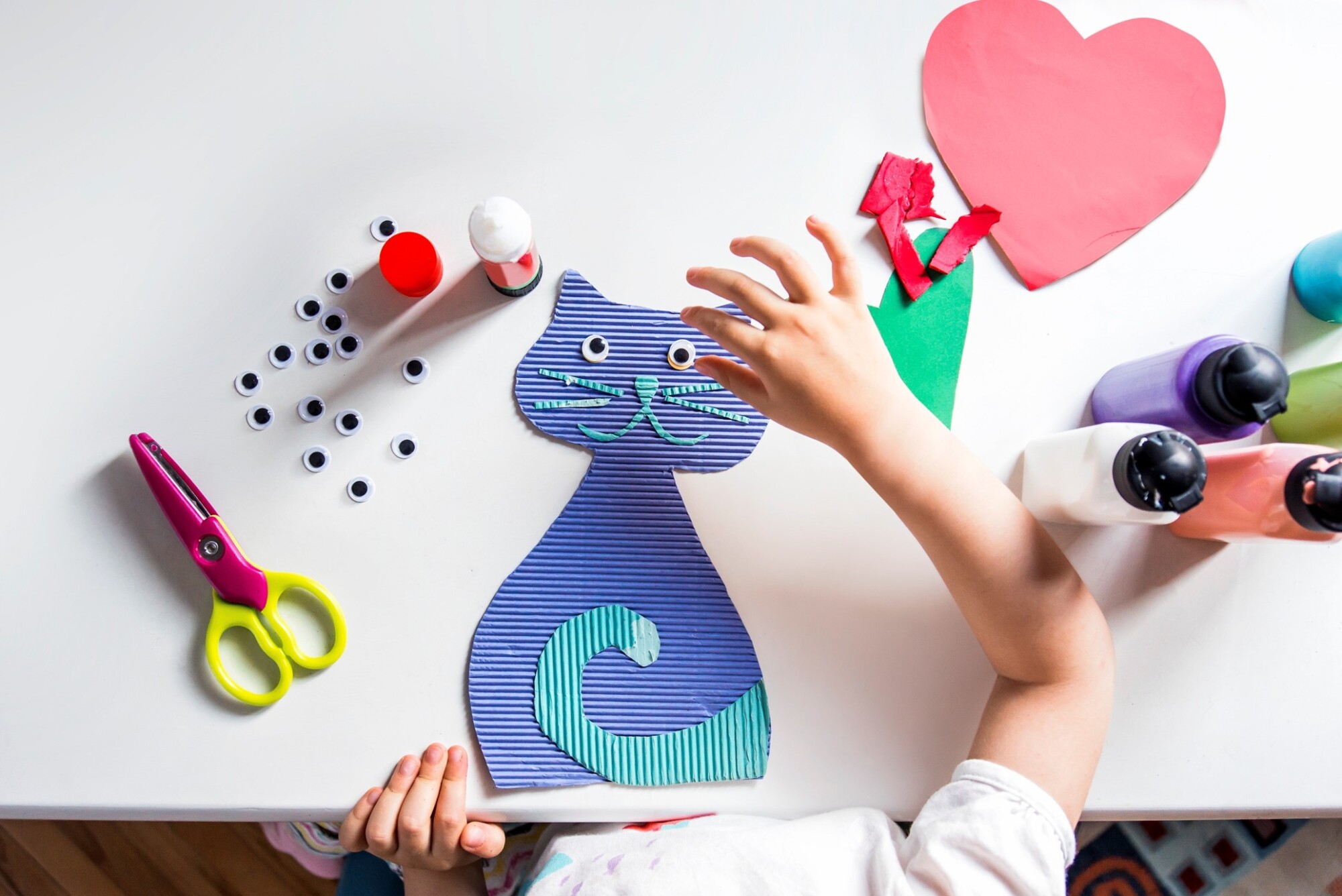 Lapsen kädet tekevät siniraidallista paperikissaa, jolla on kikkasilmät, ja ympärillä on askartelutarvikkeita, kuten sakset, liimaa, värillistä paperia ja maalipulloja valkoisella pöydällä.