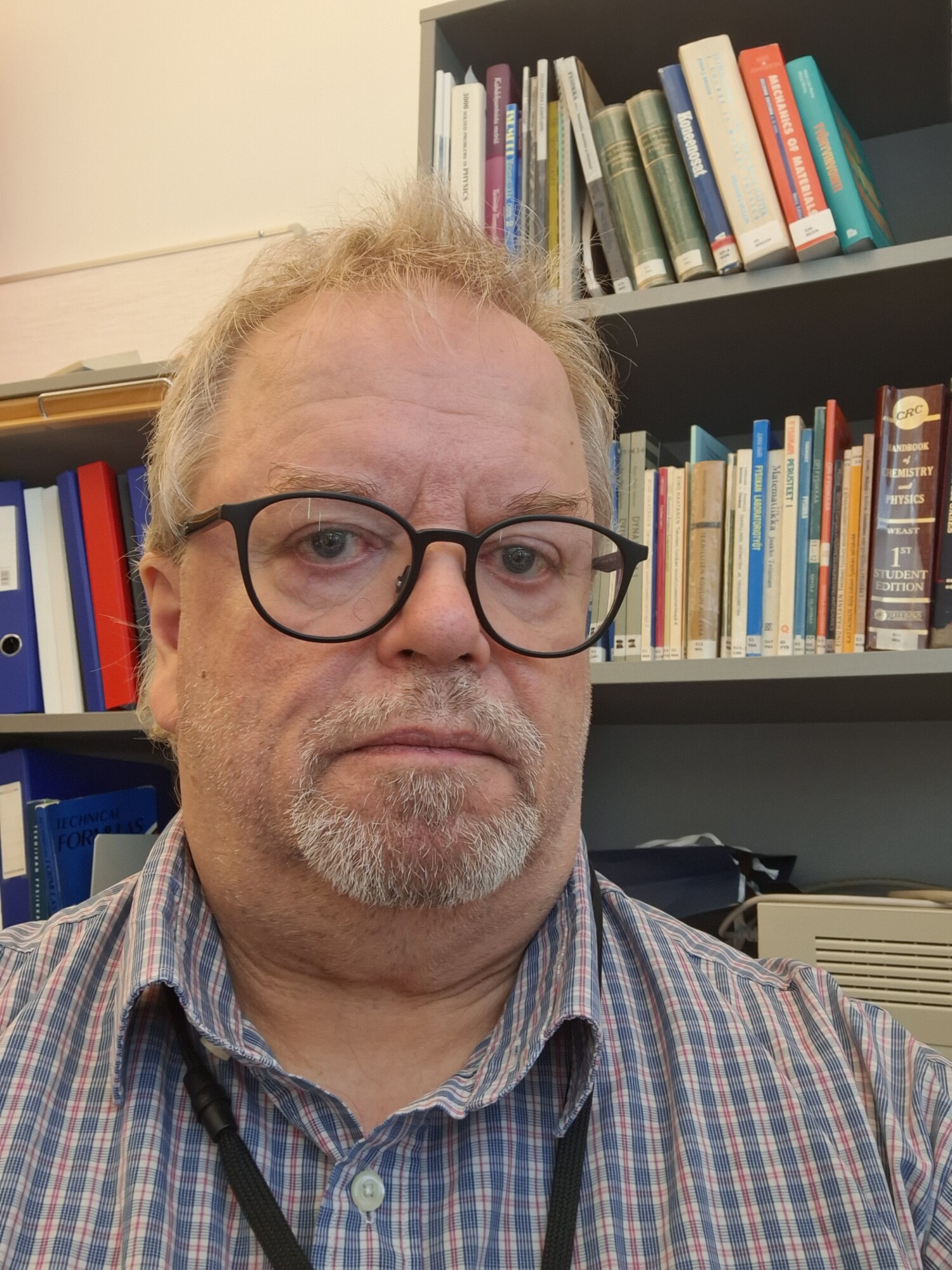 Keski-ikäinen mies, jolla on silmälasit, lyhyet vaaleat hiukset ja harmaa vuohenpukki ja jolla on ruudullinen paita, istuu kirjahyllyn edessä, joka on täynnä kirjoja ja kansioita.