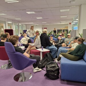 Ryhmä opiskelijoita istuu ja juttelee modernissa oleskelutilassa, jossa on violetteja tuoleja, sinisiä sohvia ja violetti matto. Jotkut rentoutuvat, toiset keskustelevat, ja reput ovat lattialla. Tunnelma vaikuttaa rennolta ja sosiaaliselta.