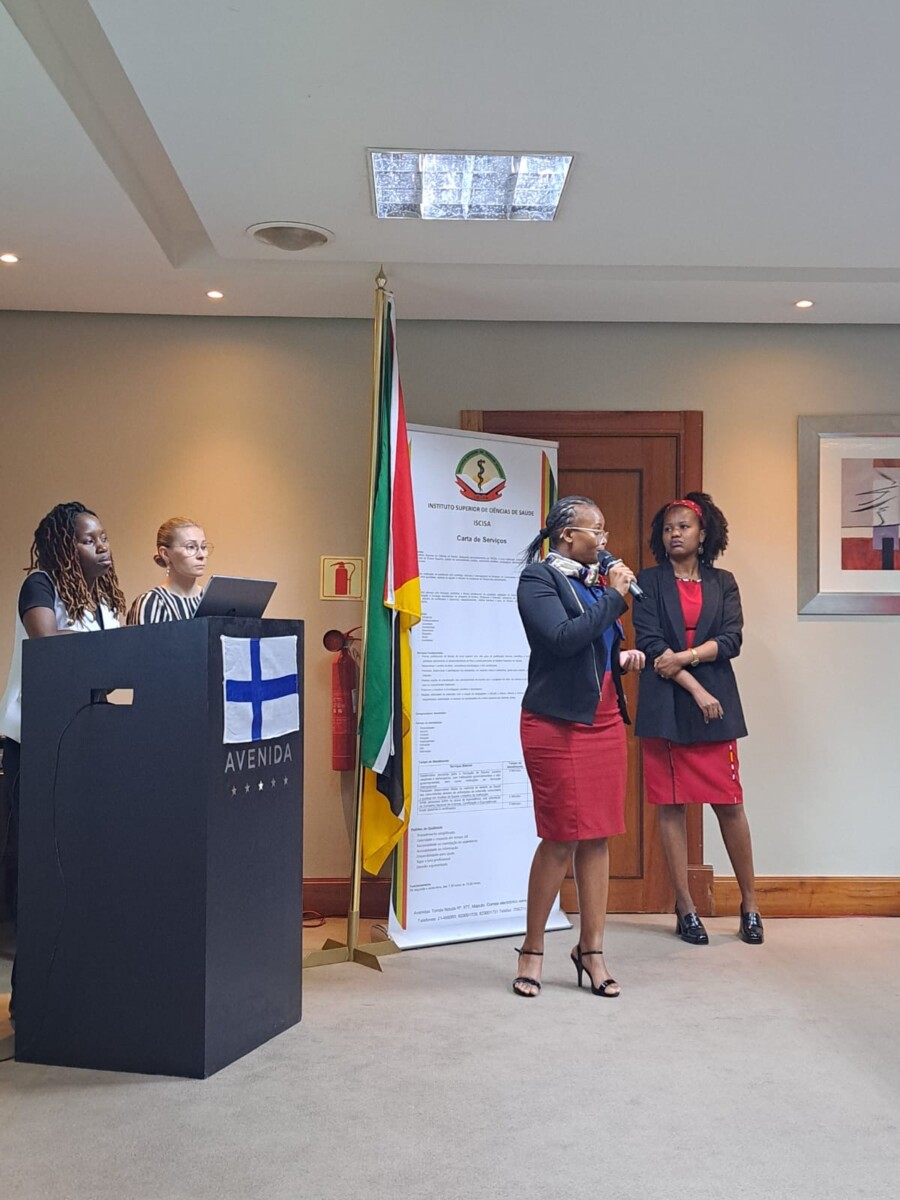 Neljä naista seisoo neuvotteluhuoneessa. Yksi nainen puhuu mikrofoniin, kun muut seisovat korokkeen lähellä, jossa on Avenida-kyltti. Taustalla näkyy lippuja ja suuri juliste.