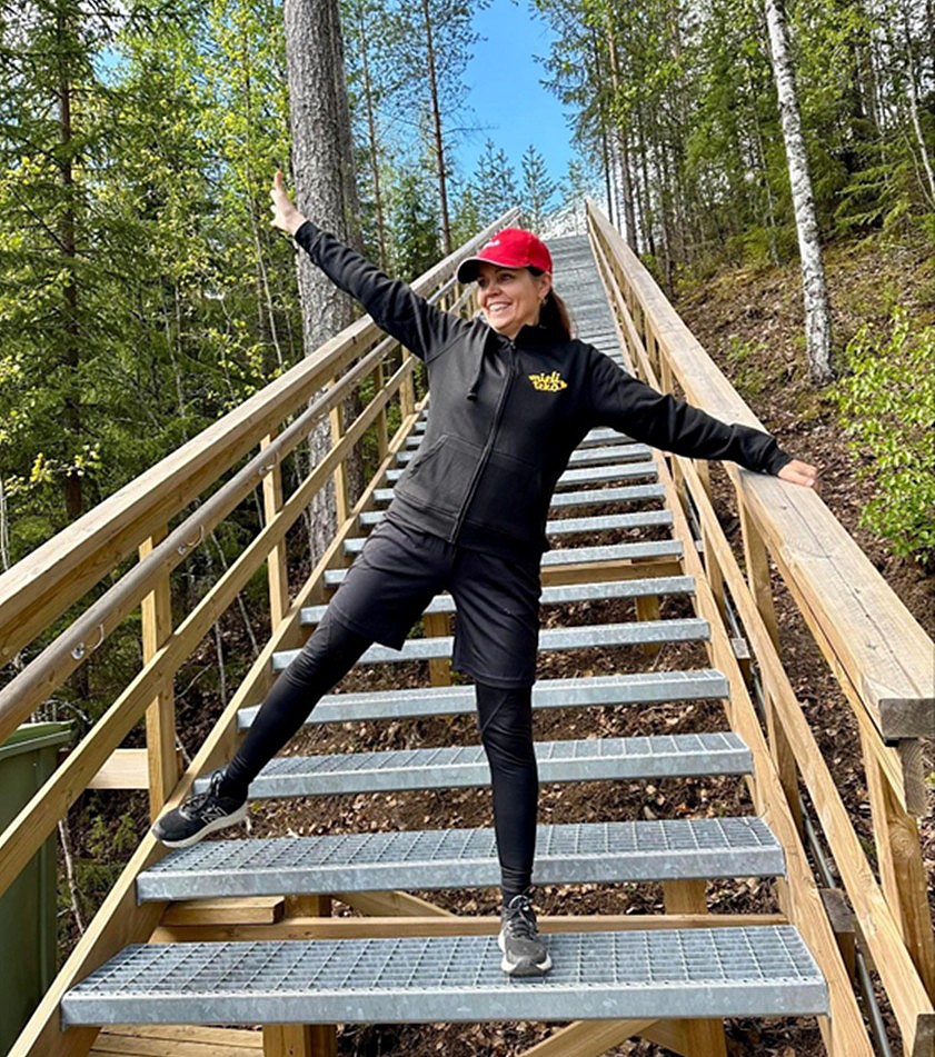 Mustiin urheiluvaatteisiin ja punaiseen lippikseen pukeutunut henkilö poseeraa leikkisästi metsäisellä alueella sijaitsevilla metalliportailla, toinen käsi koholla ja toinen jalka ojennettuna. Puut ja vehreys ympäröivät portaita.