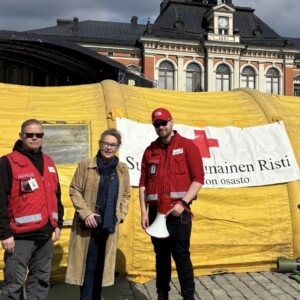 Kolme ihmistä seisoo keltaisen teltan edessä, jossa on Punaisen Ristin lippu. Kahdella on punaiset liivit, ja toisella on kädessään megafoni, kun taas kolmas henkilö seisoo heidän välissään. Taustalla on historiallinen rakennus ja pilvinen taivas.