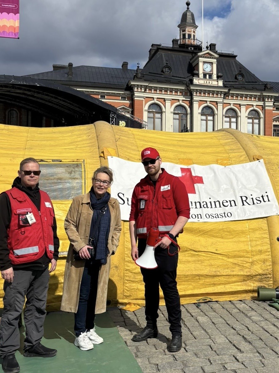 Kolme ihmistä seisoo keltaisen teltan edessä, jossa on Punaisen Ristin lippu. Kahdella on punaiset liivit, ja toisella on kädessään megafoni, kun taas kolmas henkilö seisoo heidän välissään. Taustalla on historiallinen rakennus ja pilvinen taivas.