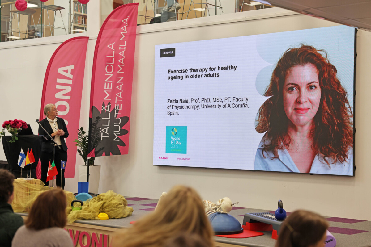 Puhuja seisoo korokkeella yleisön edessä, ja näytöllä näkyy Zélia Naian esitys "Exercise therapy for healthy ageing in older adults", jossa on hänen kuvansa ja valtakirjansa.