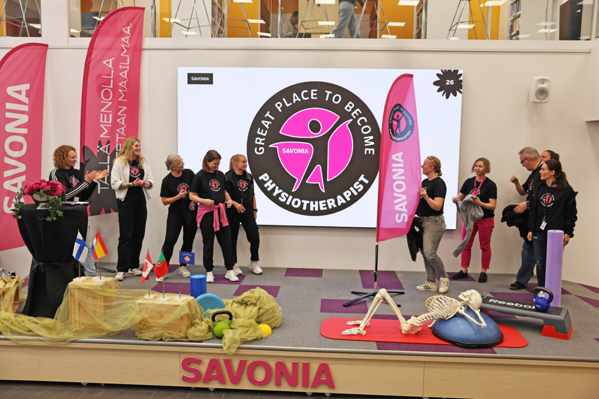Ryhmä mustiin paitoihin pukeutuneita ihmisiä seisoo lavalla, jossa on fitnessrekvisiittaa, lippuja ja luurankomalli. Heidän takanaan olevalla suurella näytöllä lukee Great Place to Become Physiotherapist, Savonia. Vaaleanpunaisissa banderolleissa lukee SAVONIA.