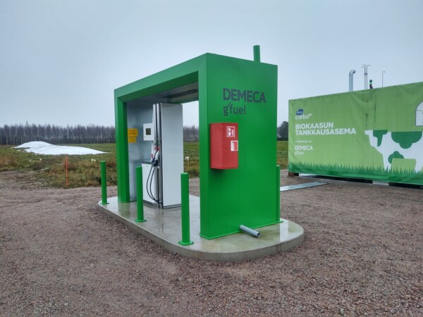 Pieni, vihreä biokaasun tankkausasema seisoo ulkona soralla, ja siihen on kiinnitetty polttoainepumppu ja punainen sammutuslaatikko. Taustalla on vihreä kontti, jossa on tietoa biokaasusta. Taivas on pilvinen.