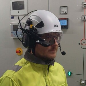 Henkilö, jolla on valkoinen Petzl-kypärä, suojalasit ja näkyvyystakki sekä kuulokkeet ja älylasit, seisoo kytkimillä ja digitaalinäytöillä varustettujen ohjauspaneelien edessä.