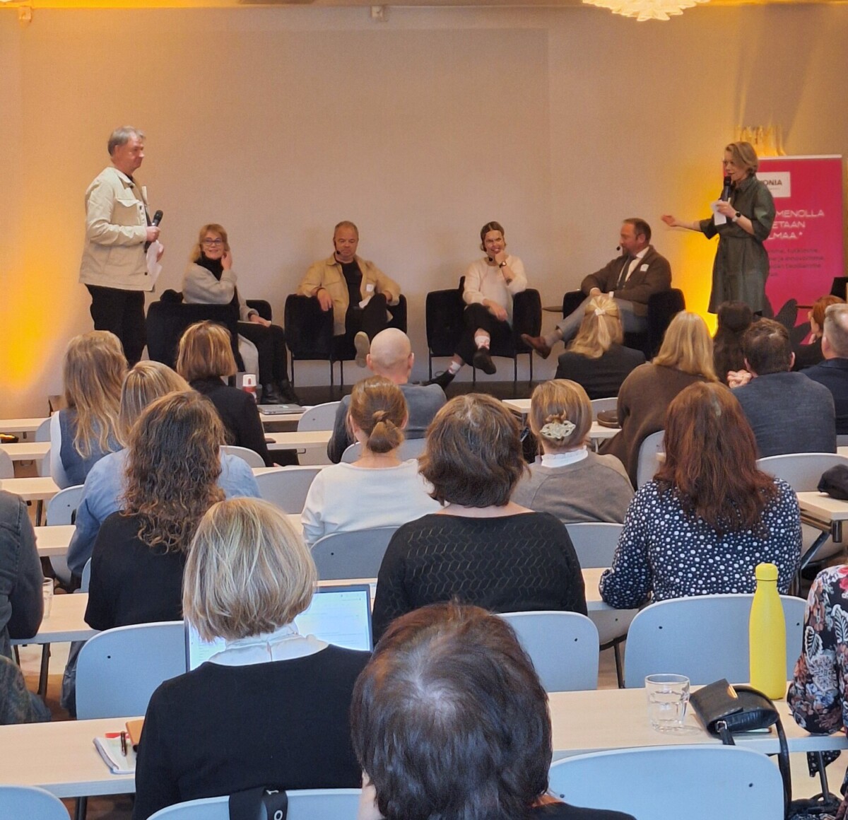 Paneelikeskustelu käydään kokoushuoneessa. Viisi henkilöä istuu lavalla, kun taas yksi henkilö seisoo ja puhuu mikrofonin avulla. Yleisö istuu pöydissä, jotka ovat lavaa vastapäätä, ja osa heistä tekee muistiinpanoja.