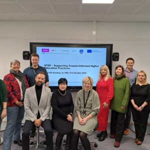 Kaksitoista ihmistä poseeraa ja hymyilee suuren näytön edessä, jossa näkyy esitys "STEP - Supporting Trauma-Informed Higher Education Practices" modernissa luokkahuoneessa tai kokoushuoneessa.