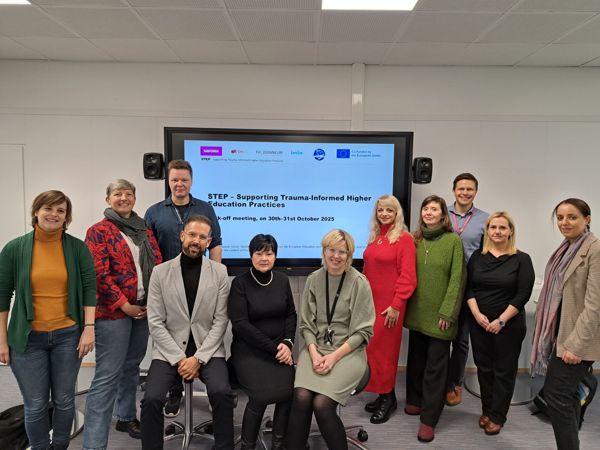 Kaksitoista ihmistä poseeraa ja hymyilee suuren näytön edessä, jossa näkyy esitys "STEP - Supporting Trauma-Informed Higher Education Practices" modernissa luokkahuoneessa tai kokoushuoneessa.