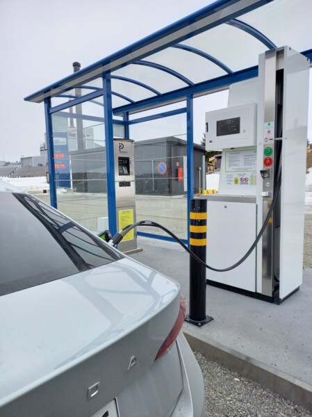 Hopeanvärinen sähköauto latautuu ulkolatausasemalla sinisen katoksen alla. Latauskaapeli on kytketty autoon, ja taustalla näkyy rakennuksia.