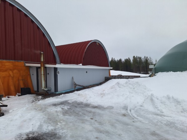 Lumipeitteinen alue, jossa on kaksi suurta kaarevakattoista rakennusta, toinen punainen, jossa on metallinen savupiippu, ja toinen vihreä, puiden ympäröimänä pilvisen taivaan alla. Punaisen rakennuksen kyljessä näkyy numero 15.