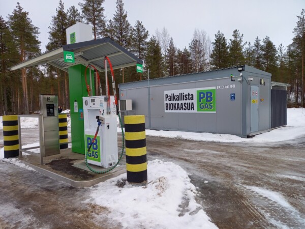 Biokaasun tankkausasema pumppuineen ja letkuineen seisoo Paikallista Biokaasua ja PB GAS:ia kuvaavan harmaan rakennuksen vieressä lumisella maalla, mäntyjen ympäröimänä pilvisen taivaan alla.