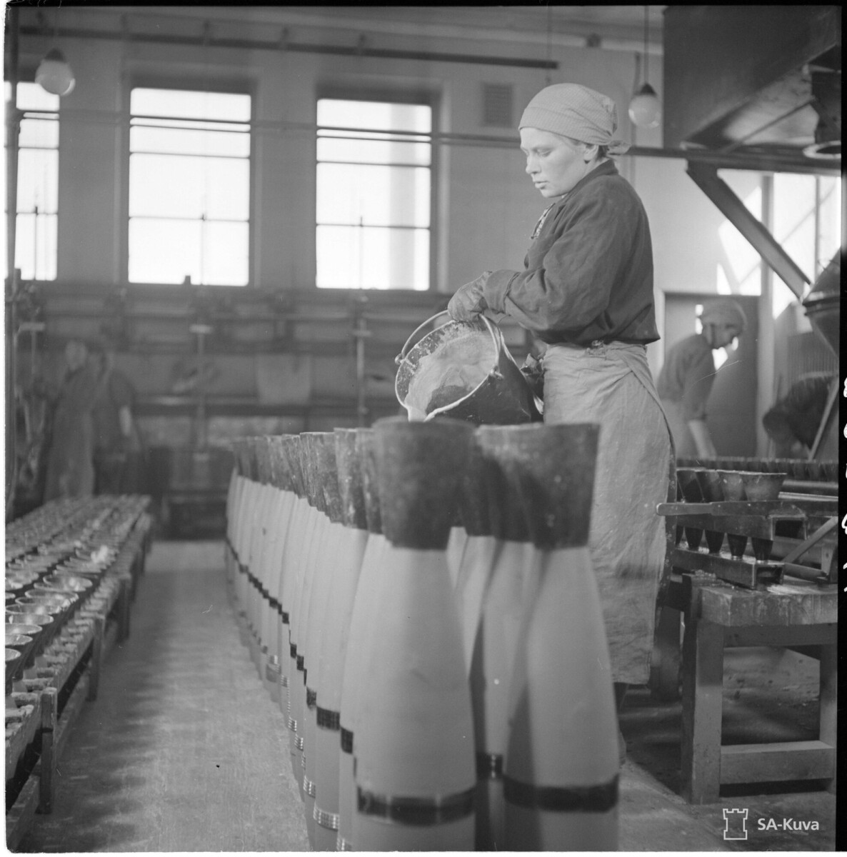 Työvaatteisiin ja huiviin pukeutunut nainen kaataa materiaalia ämpäristä tykistön kranaatteihin, jotka ovat rivissä tehtaan pöydällä, ja taustalla on muita työntekijöitä. Mustavalkoinen kuva.