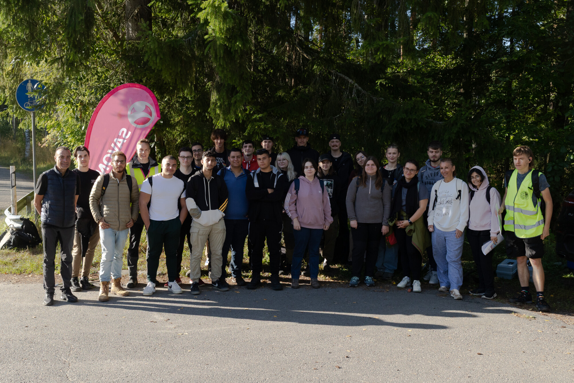 Noin 25 hengen ryhmä, joka seisoo yhdessä ulkona aurinkoisena päivänä puiden edessä ja poseeraa valokuvaa varten. Joillakin on rennot vaatteet, muutamilla on reput, ja yhdellä henkilöllä on keltainen heijastinliivi. Vasemmalla on vaaleanpunainen lippu.