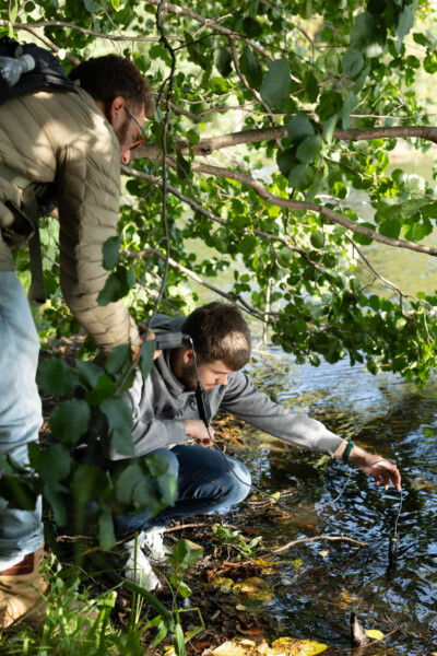 Kaksi ihmistä joen rannalla, toinen kyyristyy ja kurkottaa veteen kepin kanssa, toinen seisoo lähellä takki päällä, molempia ympäröivät vihreät lehtien oksat.