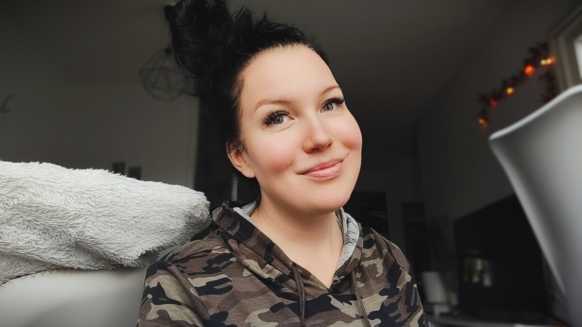 Nainen, jolla on pitkät tummat hiukset sotkuisessa pussissa, hymyilee kameralle. Hänellä on yllään maastohuppari, ja hän on sisätiloissa, joissa on viihtyisä, pehmeästi valaistu tausta.