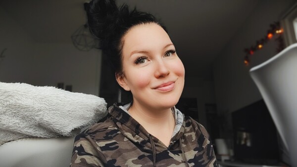 Nainen, jolla on pitkät tummat hiukset sotkuisessa pussissa, hymyilee kameralle. Hänellä on yllään maastohuppari, ja hän on sisätiloissa, joissa on viihtyisä, pehmeästi valaistu tausta.