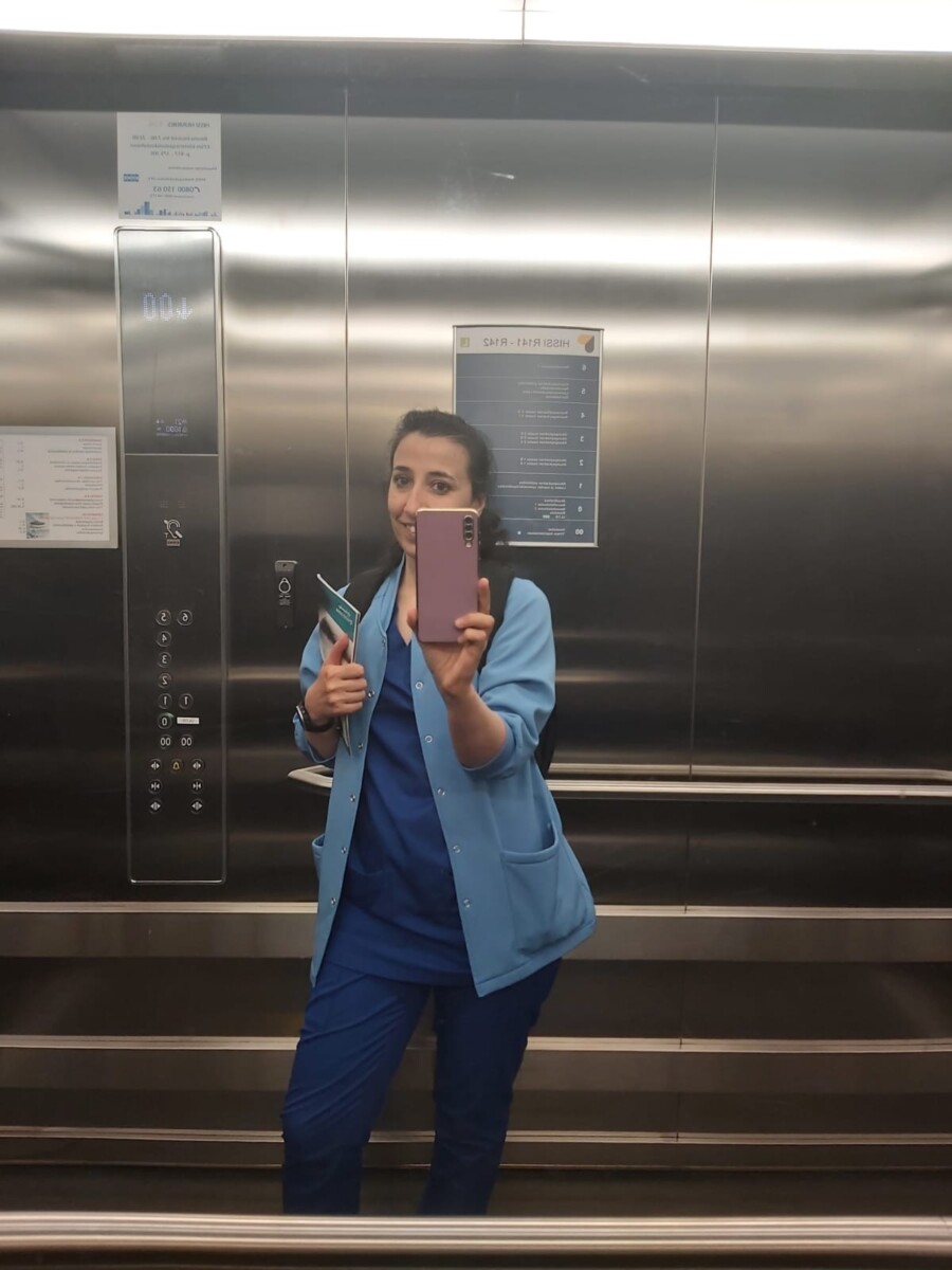 Sinisiin hoitohousuihin ja vaaleansiniseen takkiin pukeutunut nainen seisoo hississä, hymyilee ja ottaa älypuhelimellaan selfien peiliin. Hississä on metalliset seinät ja näkyvät ohjauspaneelit.