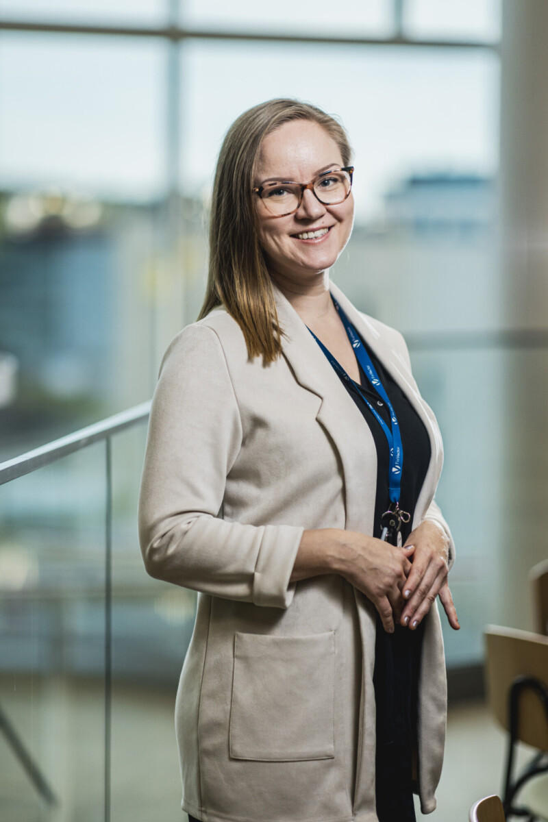 Silmälasipäinen nainen, jolla on suorat, olkapään pituiset hiukset, seisoo sisätiloissa ja hymyilee kameralle. Hänellä on yllään vaaleanbeige bleiseri tumman topin ja sinisen kaulaliinan päällä, ja hänen takanaan on sumea ikkunallinen tausta.