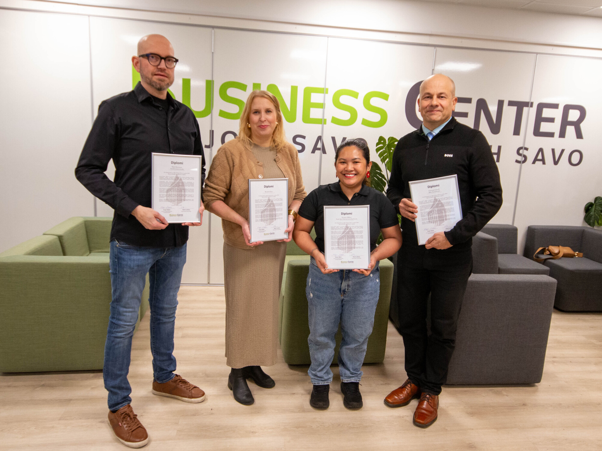 Neljä ihmistä seisoo sisätiloissa pitelemässä todistuksia ja hymyilemässä kameralle. Heidän takanaan on seinällä teksti Business Center Pohjois-Savo. Taustalla näkyy kaksi sohvaa ja pieni pöytä.