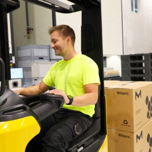 Kirkkaankeltaiseen paitaan pukeutunut mies käyttää varastossa trukkia ja siirtää hymyillen laatikoita. Taustalla näkyy pinottuja kuormalavoja ja varastohyllyjä.