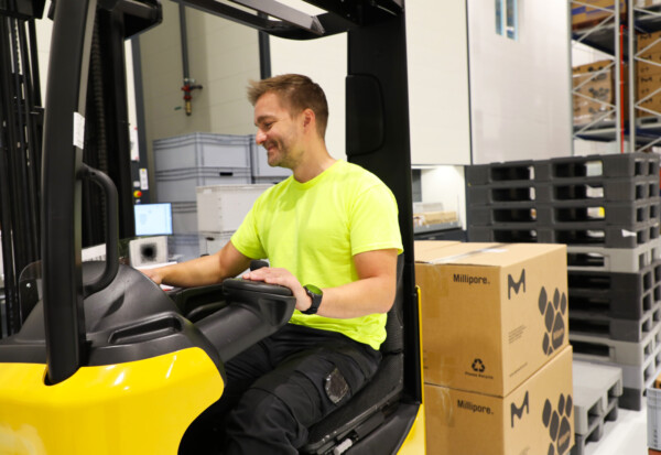 Kirkkaankeltaiseen paitaan pukeutunut mies käyttää varastossa trukkia ja siirtää hymyillen laatikoita. Taustalla näkyy pinottuja kuormalavoja ja varastohyllyjä.
