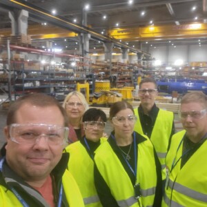 Kuusi keltaisiin turvaliiveihin ja kirkkaisiin suojalaseihin pukeutunutta ihmistä seisoo yhdessä suuressa teollisuusvarastossa, joka on täynnä metallihyllyjä, laitteita ja koneita, ja poseeraa ryhmäkuvaa varten.