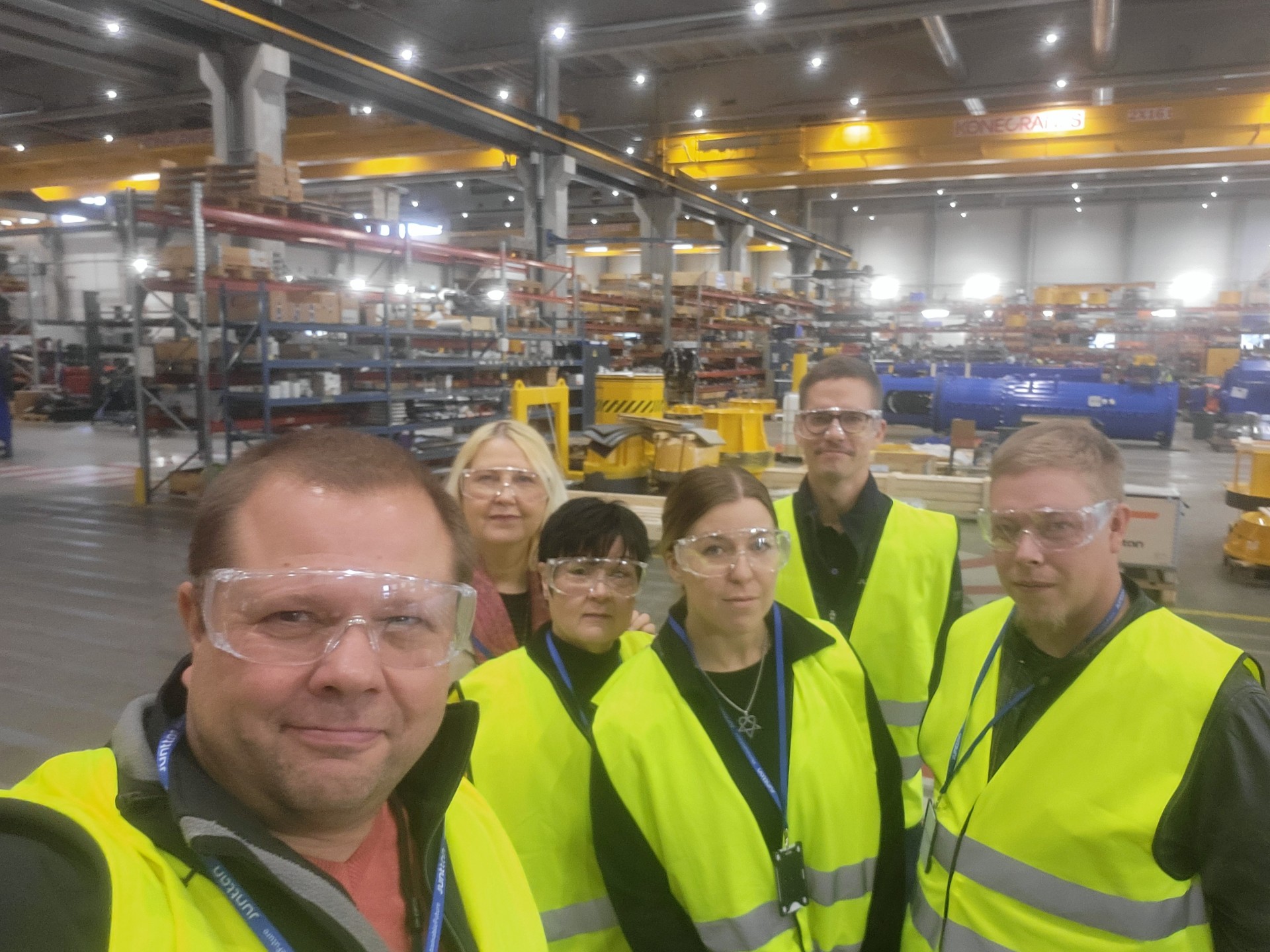 Kuusi keltaisiin turvaliiveihin ja kirkkaisiin suojalaseihin pukeutunutta ihmistä seisoo yhdessä suuressa teollisuusvarastossa, joka on täynnä metallihyllyjä, laitteita ja koneita, ja poseeraa ryhmäkuvaa varten.