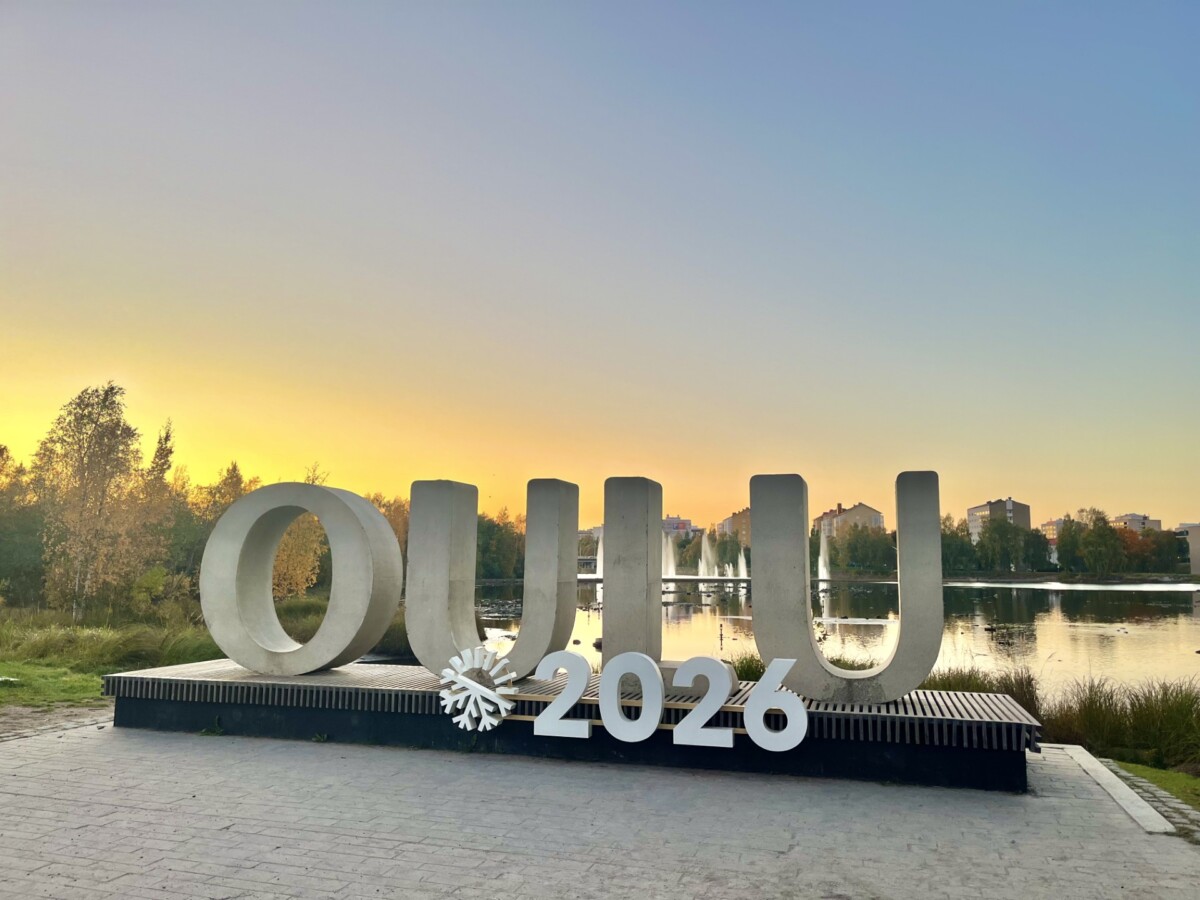 Suurilla valkoisilla kirjaimilla kirjoitetaan OULU 2026 lumihiutaleen symbolin kera, ja taustalla on seesteinen järvi ja puita värikkäällä auringonlaskun taivaalla Oulussa, Suomessa.