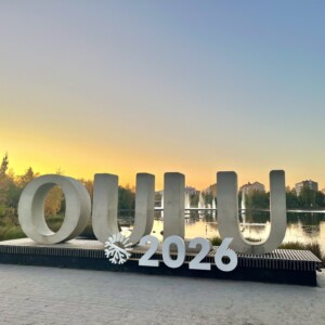 Suurilla valkoisilla kirjaimilla kirjoitetaan OULU 2026 lumihiutaleen symbolin kera, ja taustalla on seesteinen järvi ja puita värikkäällä auringonlaskun taivaalla Oulussa, Suomessa.