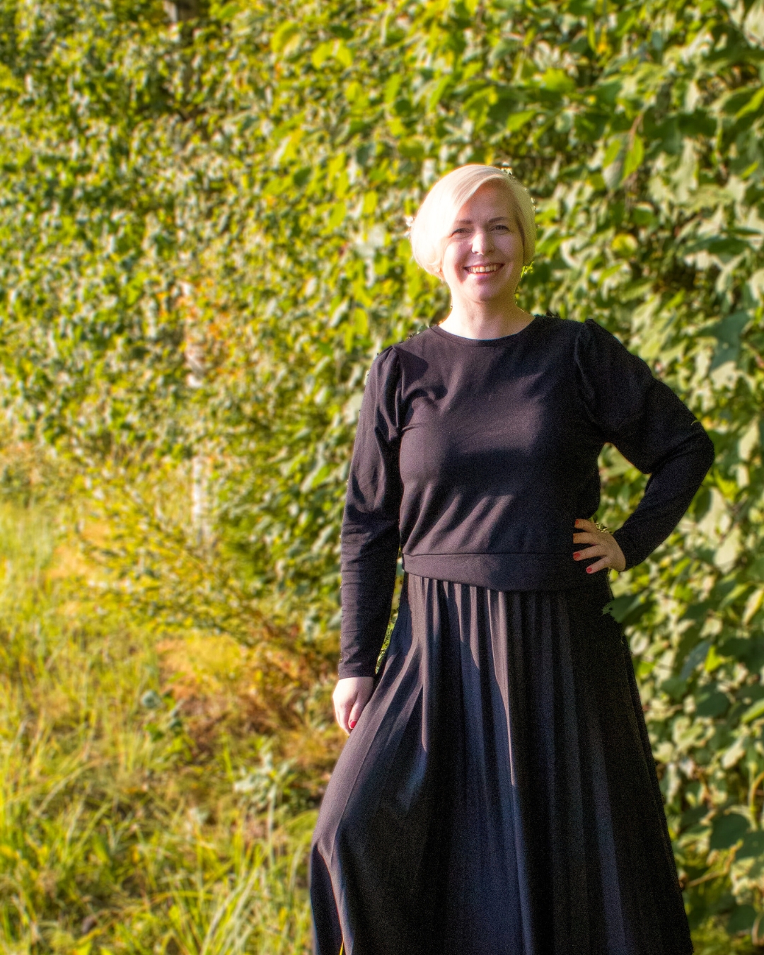 Lyhyet vaaleat hiukset omaava nainen, jolla on pitkähihainen musta toppi ja pitkä musta hame, seisoo hymyillen ulkona rehevän vihreän lehdistön edessä aurinkoisena päivänä.