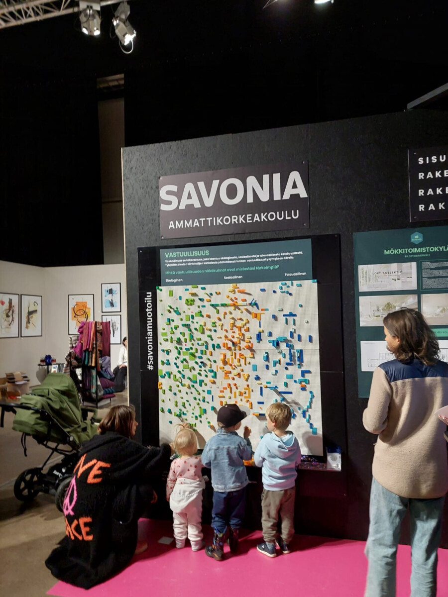 Useita pieniä lapsia seisoo värikkään interaktiivisen näyttötaulun edessä Savonia-ammattikorkeakoulun osastolla, aikuiset valvovat ja lastenrattaat ovat lähellä. Paikka on sisätiloissa ja seinällä on informatiivisia julisteita.
